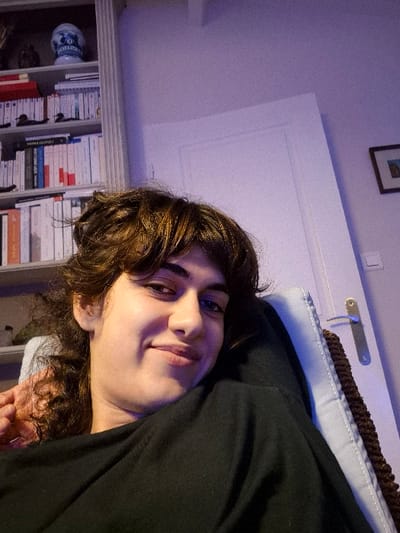 An individual with medium-length, wavy brown hair and bangs is depicted in a head-and-shoulders to upper-torso view, looking directly at the camera. The subject has fair skin, dark eyes, and a slight upward curve to the right side of the closed mouth, indicating a smile. They are wearing a plain black long-sleeved garment. The individual is seated or reclined in a light-colored upholstered chair, partially covered by a brown knitted throw or blanket.
To the left of the subject, a white multi-shelf bookcase is visible, stocked with numerous books displaying spines of various colors, predominantly white, red, and black. A blue and white ceramic vessel and other small, indistinct objects are placed on the top shelf. To the right of the subject, a white interior door with a silver-colored lever handle is visible. A framed picture hangs on the wall above and to the right of the door handle. The room walls are painted a light lavender or pale purple hue, under interior artificial lighting conditions. This indoor scene is located in Châtelaillon-Plage, France.