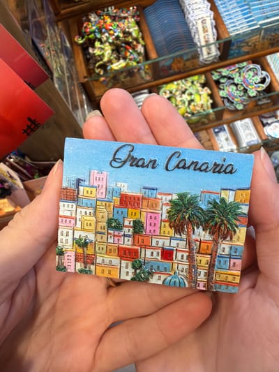 The image displays a rectangular souvenir magnet, approximately 10-12 cm in width, held horizontally by a person's light-skinned hands in an indoor retail setting. The magnet features the embossed dark brown text "Gran Canaria" in a cursive font, set against a light blue background representing the sky. Below the text, a vibrant, three-dimensional relief depicts a dense, colorful townscape with numerous multi-story buildings in varying hues including pink, yellow, blue, red, orange, and white, suggesting a hillside settlement. Three palm trees are interspersed within this townscape, with two prominent ones on the right side. A small blue dome structure is visible at the bottom center of the magnet's illustration. A gold-colored ring is present on the ring finger of the left hand. The background, out of focus, reveals wooden display shelving filled with assorted souvenir merchandise, including small colorful figurines and packaged items. The scene is consistent with a souvenir shop located in the city of Arucas, Gran Canaria, Spain.