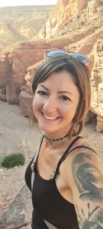 A woman with tattoos on her arm and neck is smiling at the camera. She has glitter makeup around her eyes, a nose ring, and sunglasses on her head. She's wearing a black top and a necklace. The background shows a rocky landscape. The location is identified as Hevel Eilot Regional Council, Israel.