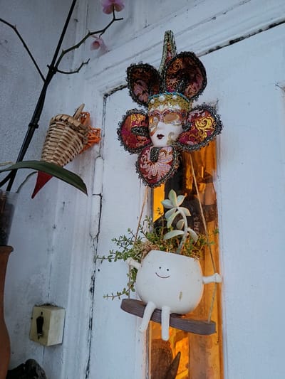 On a white, cracked, and peeling wall in Gulrypsh, Abkhazia, hangs an eclectic collection of decorations. At the top, a decorative mask with a pale face and ornate, dark red and gold headdress. Below the mask is a white, round planter with a smiling face drawn on it, filled with green plants. The planter is suspended as a swing from a small wooden plank hanging by twine. To the left of these, a small woven basket hangs on the wall. A thin branch with a few light pink orchid blossoms is seen in the upper left corner. Further to the left and below, a clay pot is partially visible, as is a vintage white electrical switch. A warm, orange glow emanates from behind the decorations, possibly indicating light from an interior space coming through a window or doorway.