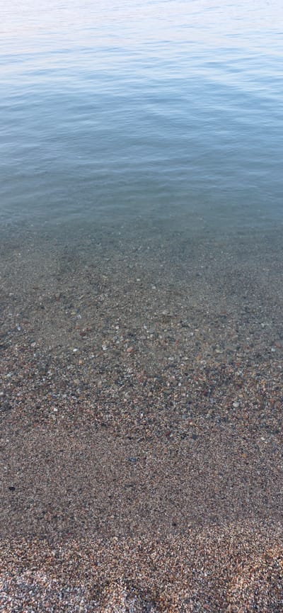 This image depicts a tranquil scene at the shore of a body of water, likely a lake or calm sea. The water occupies the upper portion of the frame, exhibiting gentle ripples and a gradient of light blue hues that suggest clear, shallow depths. The bottom half of the image showcases a beach composed of fine pebbles and sand, transitioning from wet, darker stones submerged at the water's edge to drier, lighter colored material further inland. The overall impression is one of natural serenity, devoid of any human presence or artificial elements. The location is identified as Boz Beshik, Kyrgyzstan.