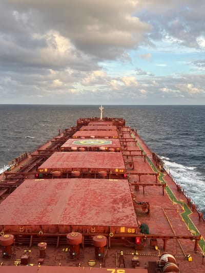 An eye-level, full shot captures a stern view of a large cargo ship at sea, its red deck stretching towards the horizon. The ocean, a deep blue-gray, is choppy with whitecaps. Above, a dramatic sky filled with a mixture of dark, heavy clouds and patches of lighter, diffused sunlight creates a moody atmosphere. The ship's deck is segmented by large, red, metal hatch covers, some marked with yellow lettering and numbers like "5P," "6P," "5S," and "6S." A helipad, marked with a white "H" and a green circle, is visible on one of the central hatch covers. Various pipes, railings, and circular ventilation covers are also present on the deck. The scene is devoid of people. The location is Unknown, Papua New Guinea.