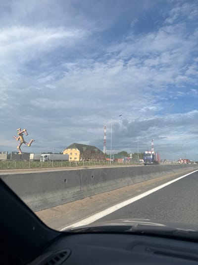 The image, taken in Beloborodovo, Russia, captures a bright daytime scene from the perspective of a vehicle, looking out onto a road and adjacent industrial landscape. On the right side, a paved road with a solid white line runs alongside a concrete barrier or curb. A dark grey car interior element is visible in the bottom right corner, indicating the viewpoint from inside a vehicle.

Beyond the road and barrier, the scene unfolds with a mix of industrial and commercial elements under a partly cloudy blue sky. Several tall structures with distinctive red and white stripes, resembling communication towers or industrial stacks, pierce the skyline in the mid-ground and background. A large, multi-story yellow-beige building with a green roof, possibly a warehouse or commercial facility, is also prominent. Scattered streetlights line the roadside.

In the distance, various trucks, including a blue truck and a white semi-trailer, are visible, suggesting an active industrial or transport area. A particularly notable object is a large, yellow and brown, multi-limbed inflatable tube figure, resembling a stylized "wacky waving inflatable arm flailing tube man," positioned in the lower-left area of the frame. It appears to be standing or "flying" near the road. The weather is clear to partly cloudy, with ample sunlight illuminating the scene. No discernible text is present in the image.