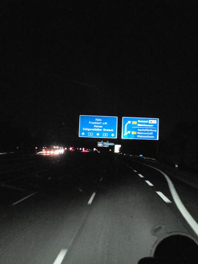 The image captures a nighttime view of a German Autobahn, likely near Kleinostheim. Two blue highway signs are prominently displayed, directing traffic. The sign on the left indicates routes to "Köln," "Frankfurt a.M.," "Hanau," and "Seligenstädter Dreieck," with directional arrows and lane numbers. The sign on the right points towards "Stadtteil," "Mainhausen," "Aschaffenburg," and "Mainaschaff Kleinostheim," also featuring directional arrows. The road ahead is illuminated by the headlights of oncoming traffic, creating streaks of light, and the white lane markings are visible. The environment is dark, consistent with nighttime driving. There are no visible people or specific interactions depicted.