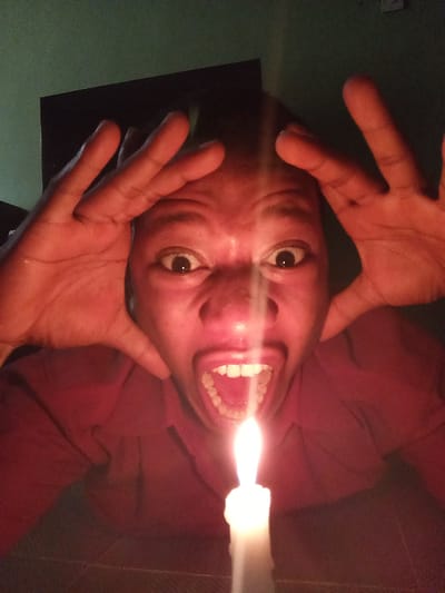 A medium, low-angle shot shows a Black person with light-brown skin, large eyes and open mouth, hands raised on both sides of the head, and illuminated by a white candle placed close to the face and directly below the mouth. The environment is dimly lit, and the candle casts shadows across the subject's face and body. The background is dark and indistinct, suggesting an indoor setting. The person appears to be looking directly at the camera with an exaggerated expression. There is no clear evidence of a specific location in Oshogbo, Nigeria, or any other identifiable geographic information in the image. The primary objects are the person, their hands, and the candle. The action is the person posing with an exaggerated expression in close proximity to the candle.