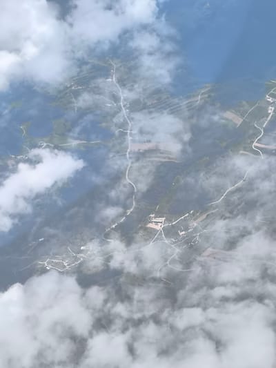 The image presents an aerial view of a landscape partially obscured by clouds. Visible features include bodies of water, likely lakes or reservoirs, agricultural fields, and forested areas interspersed with roads. In the bottom portion of the image, a cluster of buildings suggests a settlement. Given the prompt, this likely represents a region near Fischamend, Austria. No people or specific actions are discernible. The environment is rural with a mix of natural and man-made elements.