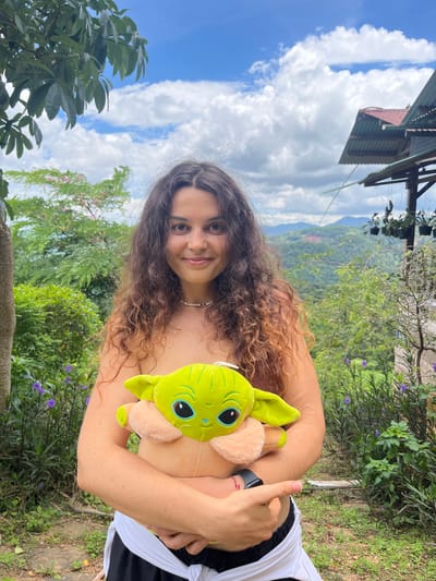 A young woman with long, curly brown hair is holding a plush toy of the character Grogu. She is wearing a white beaded necklace and a black smartwatch on her left wrist. She is wearing white fabric tied around her waist over black pants. She is standing outdoors in a lush, green environment with a mountainous landscape in the background under a cloudy sky. The location appears to be a rural hillside. There is a building with a tin roof visible in the upper right of the image, and various plants and trees surround the woman.