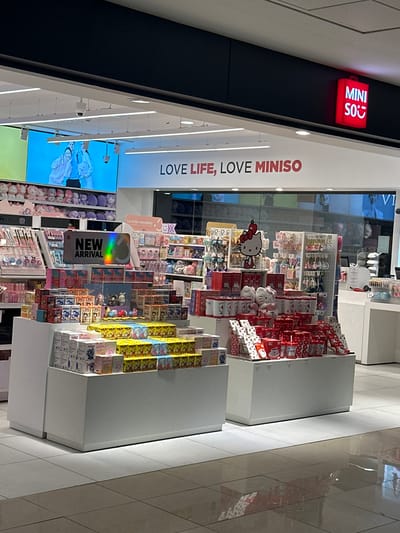 This is an interior view of a MINISO store, a retail establishment that offers a variety of lifestyle products. The store is identified by a red sign with the brand name "MINISO" displayed on a black background. The store's slogan, "LOVE LIFE, LOVE MINISO," is prominently featured on a white wall. The display cases are stocked with items such as Hello Kitty merchandise, plush toys, and blind boxes, all of which are neatly arranged. A sign reading "NEW ARRIVAL" indicates the presence of new products. The location of the MINISO store is in the city of Pudahuel, Chile, based on the context and visual evidence. The background features various items for sale and the overall environment is brightly lit and organized.