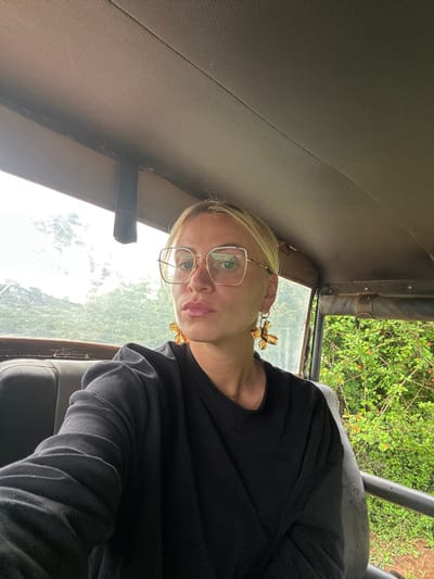 A forensic examination of the image reveals a fair-skinned individual, identified as a woman, seated inside what appears to be an open-sided safari-style vehicle. The setting is Situlpawwa, Sri Lanka.

The woman is positioned centrally, facing directly towards the camera. She possesses blonde hair, pulled back, and wears large, gold-framed, square-shaped eyeglasses. Her lips are slightly parted, exhibiting a light pink or natural tone. Prominent, amber-colored, multi-petal flower-shaped earrings with silver-toned findings are visible on both ears. She is dressed in a dark, possibly black, long-sleeved garment, consistent with a sweatshirt or thick long-sleeved top. Her left arm is partially visible, bent at the elbow, suggesting she may be holding the camera or phone for a self-portrait.

The interior of the vehicle features a light brown or tan padded roof liner, with visible stitching and a dark strap hanging down on the left side. A dark gray or black seat back is partially visible behind and to the left of the woman. To the right, through an open side of the vehicle, lush green vegetation is distinctly visible, including branches with small reddish-orange elements, possibly berries or flowers. The overall impression is an outdoor, natural environment, consistent with a safari or nature reserve setting.
