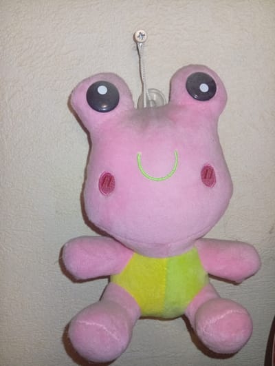 A pink plush frog toy with large black eyes, a green stitched smile, and two pink oval blush marks on its cheeks is hanging from a screw in a textured, light-colored wall. The frog is wearing a yellow and light green patched belly. The toy is suspended by a white string attached to a clear plastic suction cup, which is itself attached to the screw. The background is a wall with a rough, matte texture, suggesting it is an interior surface. The lighting appears to be artificial and uneven, casting subtle shadows. This image was taken in Iskitim, Russia.