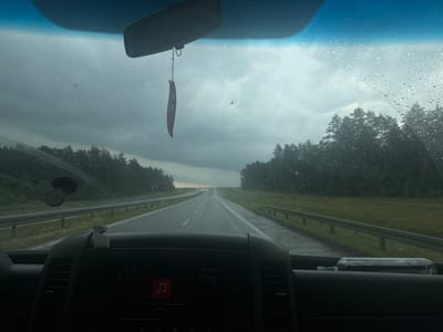 The image is taken from inside a vehicle, looking out through the rain-streaked windshield. The view is of a highway with trees lining both sides. The sky is overcast, suggesting a cloudy or rainy day. In the foreground, the dashboard of the vehicle is visible, with a dark screen displaying what appears to be a music player interface. A car air freshener hangs from the rearview mirror, and a suction cup mount is attached to the windshield. A glass bottle lies on the dashboard. The overall impression is a journey on a highway in potentially inclement weather. The location is Jarnuty, Poland.