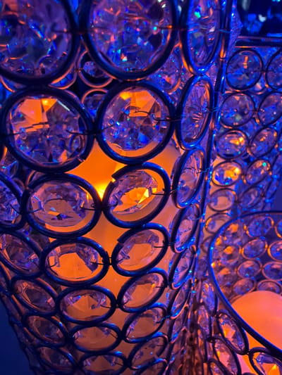 This close-up image depicts a segment of a decorative object, likely a lamp or candle holder, composed of numerous clear, faceted crystals individually set within dark metallic rings. The crystals are arranged in a tessellated pattern, forming a curved, possibly cylindrical structure. An internal, warm orange light source illuminates the object's core, casting an amber glow through the crystal facets. An external, cool blue light source illuminates the outer surfaces of the crystals and the surrounding environment, creating refractions and reflections displaying both orange and blue hues across the crystal surfaces. A portion of a second similar object is visible to the right, also illuminated. The background is dark and indistinct, suggesting an indoor setting. This scene is located in Long Beach, United States of America.