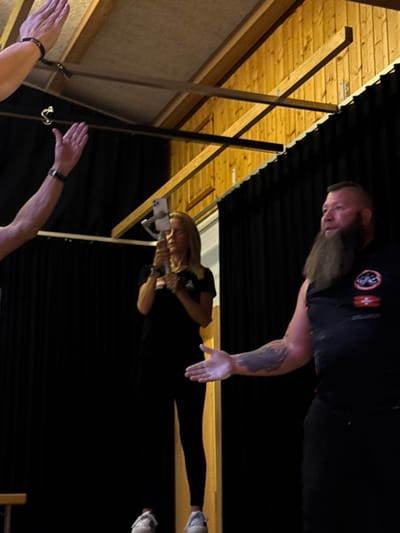 The image depicts an indoor scene in Reisenberg, Austria, featuring three individuals and a recording setup, set against a backdrop of black curtains and a wooden ceiling.
In the foreground, on the left, two arms are raised, seemingly in a gesture of waving or applause. The upper arm wears a dark wristband, and the lower arm displays a dark wristband or watch. To the right of center, a blonde woman, facing towards the right, holds a white smartphone mounted on a grey gimbal stabilizer, suggesting video recording. She wears a black t-shirt, dark leggings, and white sneakers. On the far right, a bearded man with a dark mohawk-style haircut faces left, extending his right arm outwards with an open palm. He wears a dark sleeveless top featuring a patch with a red and white cross, indicative of the Swiss flag, and other symbols on the left chest. A tattoo is visible on his right forearm.
The background consists primarily of dark black stage curtains. Above, a wooden slatted ceiling or wall section with exposed wooden beams is visible, with some metal hardware attached. Artificial lighting illuminates the subjects.