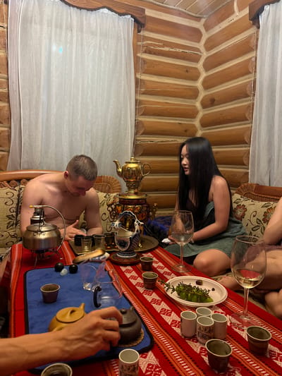 The image, rotated 90 degrees clockwise, depicts an indoor gathering within a rustic log cabin, possibly a tea ceremony. Wooden log walls and a ceiling form the background on the left and top, with white sheer curtains visible on the bottom left.

Two individuals are prominent: a woman with long dark hair, wearing a dark top, is seated in the upper-middle, looking down towards the table. Below her, a shirtless man is seated, turned towards the right, engaged with a silver kettle placed on a small burner. A third person's hand is visible on the far right, actively pouring from a terracotta-colored teapot into a small cup. Other individuals are partially visible, suggesting a larger group.

The central and right areas of the image feature a table covered with a red and white patterned tablecloth and a blue mat. The table is richly appointed with several small ceramic cups, wine glasses, and a white platter holding green grapes and dark berries. Various tea-making implements are present, including two distinct samovars—one brass and one with blue and white patterns—along with multiple teapots and small dark stones, indicating a comprehensive tea service.

The setting suggests a warm, intimate atmosphere, characteristic of a log cabin. The indoor lighting implies an evening or nighttime gathering. No visible text is present in the image.