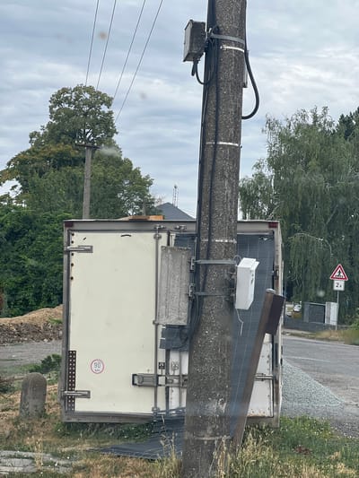 The image shows a utility pole with electrical equipment and cables attached. A refrigerated trailer is parked next to the pole. In the background, there are trees, a road, and a pedestrian crossing sign. The scene is located in Újezd u Brna, Czechia. The overall environment appears to be rural or suburban, with a mix of natural elements and man-made structures.