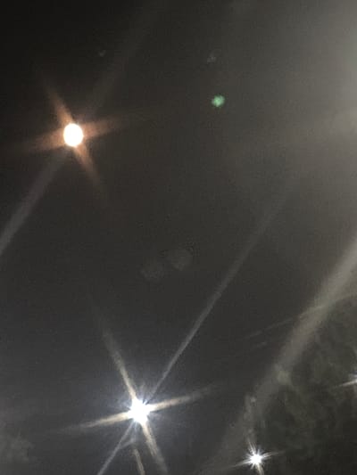 This is a dark, grainy, and slightly out-of-focus photograph taken at night, likely outdoors. The scene is dominated by the dark sky, punctuated by several bright, starburst-like light sources. The lights suggest artificial illumination, possibly from streetlights or building lights. There's a diffuse haze or fog present, which softens the edges of the lights and creates a somewhat ethereal atmosphere.
In the mid-left of the frame, there is a distinct, small, green glowing object. This object is somewhat fuzzy and appears to be isolated against the dark background. It is possibly a light source or an illuminated object of some kind. Further to the left of this green light, there's another faint, less distinct white speck.
In the lower portion of the image, there is a prominent, bright, circular light source with rays emanating outwards, creating a star-like effect. This light has a warm, yellowish-white hue. In the upper right quadrant of the image, another similar but slightly smaller bright light source is visible, also with pronounced rays. There are also faint, diagonal streaks of light across the image, suggesting reflections or light beams interacting with the atmospheric conditions or the camera lens.
The overall mood of the image is mysterious and somewhat desolate due to the darkness and the obscured nature of the scene. There are no discernible people or identifiable objects beyond the light sources. The setting appears to be an urban or semi-urban environment, given the presence of artificial lights, but the fog or haze obscures any specific location cues. The time of day is clearly night. There is no visible text in the image. The location context provided states Saran city administration, Kazakhstan, which adds a layer of understanding to the potential source of the artificial lights.