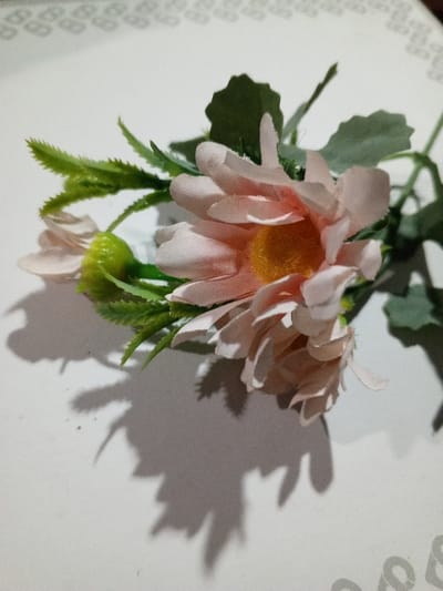 A small bouquet of artificial flowers rests on a white surface. The flowers have delicate, peach-colored petals and bright yellow centers. Green leaves and stems surround the flowers, adding texture and depth. A soft shadow is cast across the surface, highlighting the form and shape of the flowers. The background is slightly blurred with a pattern on it.