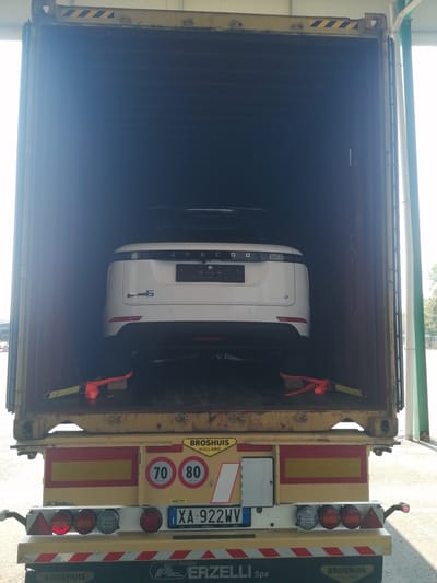 A white vehicle is parked inside an open shipping container. The vehicle is positioned at the far end of the container, facing forward. The rear of the vehicle, including its taillights and license plate, is visible. The license plate reads "XA 922WV" and has an Italian registration. The vehicle is secured with orange straps. The shipping container is mounted on a yellow truck trailer, which is also visible at the bottom of the image. The trailer has various markings, including speed limit signs (70 and 80) and the text "BROSHUIS HOLLAND". The scene appears to be outdoors, with sunlight indicating it is daytime. The location is the city of Borgolavezzaro, Italy.