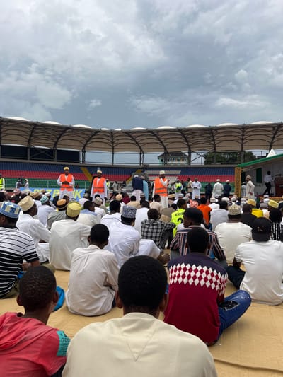 A wide-angle image captures a large gathering of individuals, predominantly male, seated on woven mats on the ground within a stadium in Zanzibar City, Tanzania. The stadium features tiered seating in red, blue, and green, and a distinctive white, arched roof structure against an overcast sky.

The seated attendees, viewed from behind, are largely dressed in traditional white Islamic robes and kufis, though some wear casual clothing including t-shirts, plaid shirts, and jeans.

In the mid-ground, several men wearing orange high-visibility vests over their attire stand facing the seated crowd, appearing to observe or manage the event. One individual in an orange vest holds a white object. A white banner with the black text "PDZ PIK" is visible behind the standing individuals.

To the right, a decorated platform with green and gold elements is set up, where several men are standing. One man on the platform addresses the gathering from a podium. A white vehicle is partially visible behind the standing group in orange vests. Trees are visible beyond the stadium structure in the distance.
