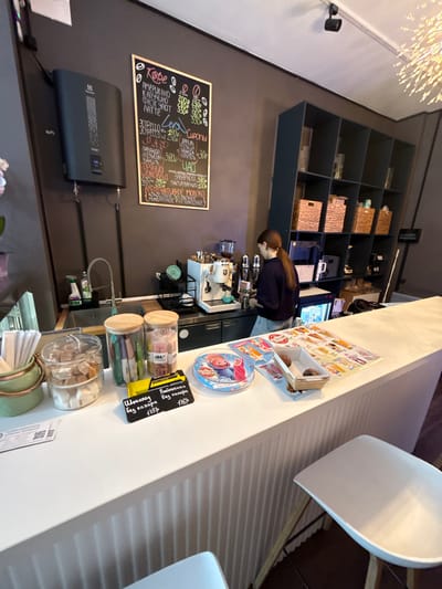 This image shows the interior of a cafe, likely in Pokrovskoe, Russia, as suggested by the location context. A young woman with dark hair tied back is visible behind a counter, appearing to be working with a coffee machine. The scene is well-lit, suggesting it's daytime.
On the wall behind the counter is a large chalkboard menu displaying various coffee drinks and their prices in Russian. Items like "АМЕРИКАНО", "КАПУЧИНО", "ФЛЭТ УАЙТ", "Эспрессо", and "ШОКОЛАД" are listed, along with a selection of "Сиропы" (syrups) such as "ЛАВАНДА" (lavender) and "Ваниль" (vanilla).
The counter itself is adorned with several glass jars containing what appear to be sugar cubes and possibly other confectionery items. There are also some packaged snacks, including a yellow packet labeled "SLIM BAR" and "Батонечки без сахара" (sugar-free bars). A small, circular item with cartoon characters, possibly a toy or a sticker, is also on the counter.
Behind the woman, shelves are stocked with various items, including woven baskets and what seem to be bottles or containers. A black electronic device, possibly a router or a modem, is mounted on the wall below the menu.
The overall atmosphere appears to be that of a casual and modern cafe, with a focus on coffee and snack offerings. The image is taken from a slightly tilted perspective.