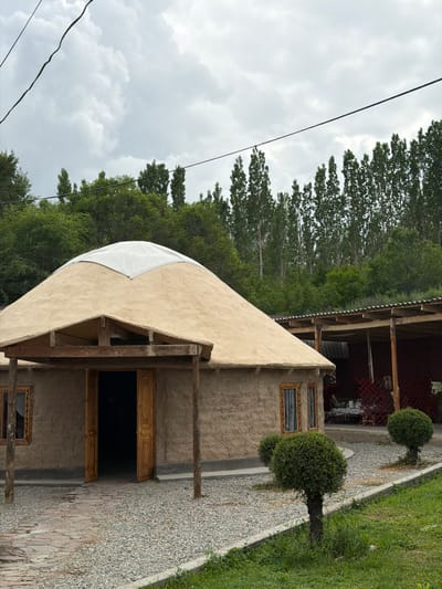 In the city of Кызыл-Туу, Kyrgyzstan, an image captures a traditional yurt with a conical roof, partially covered by a white tarp, situated in a rural setting. The yurt's walls are constructed from a rough, textured material, possibly mud or clay, with wooden window frames and a double wooden door. A covered, open-air structure with a corrugated roof is attached to the right of the yurt, containing tables and chairs, suggesting an outdoor dining or seating area. The ground in front of the yurt is covered with gravel, interspersed with a brick pathway leading to the entrance. Two small, manicured bushes flank the pathway. Behind the yurt and the adjacent structure, a dense forest of tall, slender trees stands against a cloudy sky. Overhead, electrical wires are visible against the sky. The scene appears to be peaceful and idyllic, showcasing a blend of traditional architecture and natural beauty.