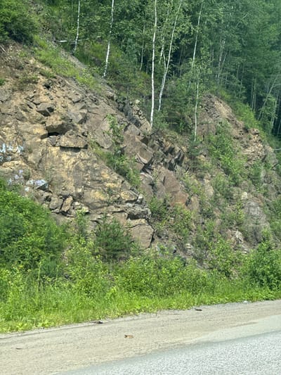 The image depicts a rocky hillside adjacent to a road, likely in Gornoural'skii, Russia. The hillside is covered in a mix of rocks, sparse vegetation, and trees. The rocks appear to be of various sizes and shapes, with some showing signs of weathering. Patches of green grass and small bushes grow between the rocks. Taller trees, some with white bark, are visible further up the slope. The road in the foreground is unpaved, with a gravel surface. The overall impression is of a natural, somewhat rugged landscape bordering a rural or undeveloped area. There are no people visible in the image.
