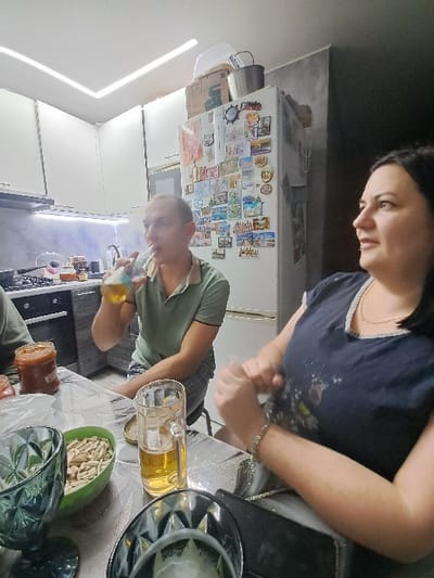 A rotated scene reveals a woman in a dark blue shirt standing behind a table laden with glasses, beer in a mug, and snacks. A man in a green shirt is present, standing near a kitchen appliance. The background includes a refrigerator covered in magnets and papers, and a kitchen with white cabinets.