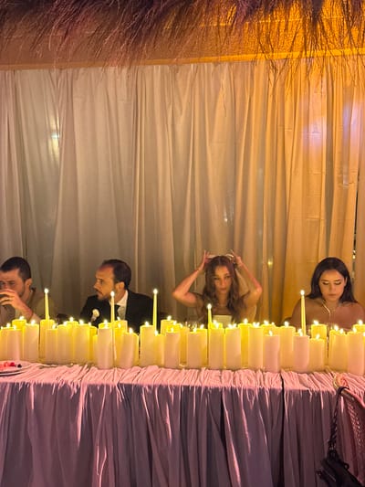 Captured in the Communauté rurale de Malicounda, Senegal, the image presents a wedding reception setting. In the frame are four individuals, two women and two men, standing around a buffet table. The table is laden with food and decorated with an array of lit candles arranged in a row along the edge. The backdrop consists of draped fabric. A floral arrangement is visible on the left side of the frame. The room has a warm tone, illuminated by the candles and overhead lighting. The subjects appear to be engaged in enjoying the event, with one woman adjusting her hair, the other standing still and the two men interacting.