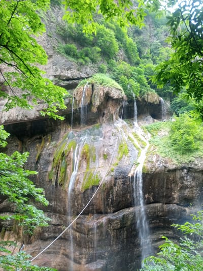 A wide shot captures a tall, multi-tiered waterfall cascading down a rugged, moss-covered rock face in Khushto-Syrt, Russia. Lush green foliage frames the scene from the top and sides. A thin white rope stretches diagonally across the lower half of the waterfall. The rock face is textured with horizontal striations and patches of green moss and lichen. The water is white and appears to be flowing rapidly. In the upper right portion of the image, a small figure, possibly a person, stands on a ledge near the top of the waterfall. The sky above is overcast.