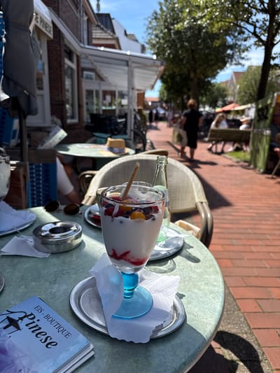 An outdoor cafe scene on the island of Langeoog, Germany, with a focus on a glass of dessert on a table. The dessert appears to be a yogurt or ice cream parfait with berries and a biscuit. Other items on the table include sunglasses, an ashtray, and a pamphlet titled "EIS - BOUTIQUE Pinese". In the background, people are seen walking and sitting along a brick-paved pathway lined with trees and buildings. The atmosphere suggests a bright, sunny day.