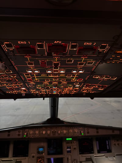 The image shows the cockpit of an airplane. The control panel is the main focus, with numerous buttons and switches illuminated in orange. Some key labels visible include "ENG 1", "ENG 2", "APU", "FIRE", "FUEL", and "HYD". The windshield and the view outside the plane can also be seen. The exterior view appears to be a tarmac or runway with a light-colored concrete surface.