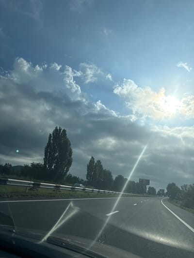 A car is driving on a highway with a guardrail on the left and trees on the right. The sky is partly cloudy with the sun shining through. The sun's rays create a lens flare across the windshield. The scene appears to be during daylight hours. The location is identified as the city of Gagarin in Armenia.