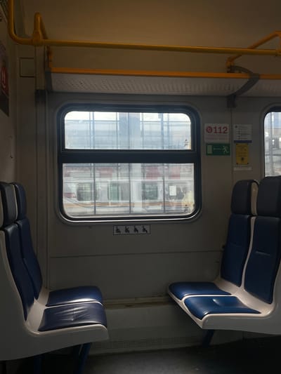 The image depicts the interior of a passenger train car, likely in Aprelevka, Russia. Two rows of blue and white seats are visible, one on the left and one on the right, facing each other across an aisle. The seats appear to be made of a synthetic material with a slightly reflective surface. The left row of seats is closer to the camera and shows two seats, while the right row has three seats visible.

A large window is situated between the two rows of seats, offering a view of another train car outside. The window is framed by the train car's interior paneling. Above the window, a yellow handrail runs horizontally along the ceiling.

Various signs are affixed to the wall next to the window. One sign, partially obscured, appears to be a red circle with the number "112" and text below it, likely indicating an emergency number. Below this is a green rectangular sign with a white arrow and a silhouette of a person running, indicating an emergency exit. Another sign, to the right, is yellow and rectangular with text in Russian and a button.

Below the window, there are four identical black icons depicting a person in a wheelchair, indicating accessible seating areas. The floor of the train car is dark and appears to be made of a durable material. The overall lighting is somewhat dim, with natural light filtering through the window and artificial lighting from within the car. The scene is static, with no people actively engaged in any discernible activity.