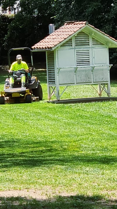 A person is operating a yellow zero-turn lawnmower on a grassy field in front of a small, open-sided white structure with a terracotta tile roof. The person is wearing a bright yellow t-shirt, a light-colored baseball cap, and gray pants with bright yellow accents. They are seated on the lawnmower, holding the steering controls. The structure has louvred panels and a counter-like area in front. The lawn is well-maintained and green, with dappled sunlight and shadows suggesting trees nearby. The background shows dark green foliage and a building with a reddish roof. The overall scene depicts groundskeeping work in an outdoor area.