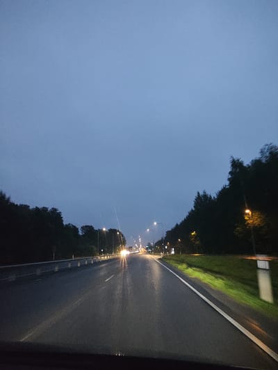 The image depicts a view from a vehicle traveling on a wet, multi-lane road at dusk or dawn. The sky is a uniform, pale blue, suggesting overcast conditions or the twilight hours. Trees line both sides of the road, forming a dense, dark border. Streetlights, some with visible glowing bulbs, are spaced along the right side of the road, illuminating the wet asphalt and the grassy verge. Headlights from oncoming traffic are visible in the distance, creating streaks of light on the road surface. A white line divides the lanes, and a metal guardrail is present on the left side. The overall impression is of a quiet, possibly rural or suburban highway, likely within or near the city of Harkujärve, Estonia, given the context. No people are visible in the frame.