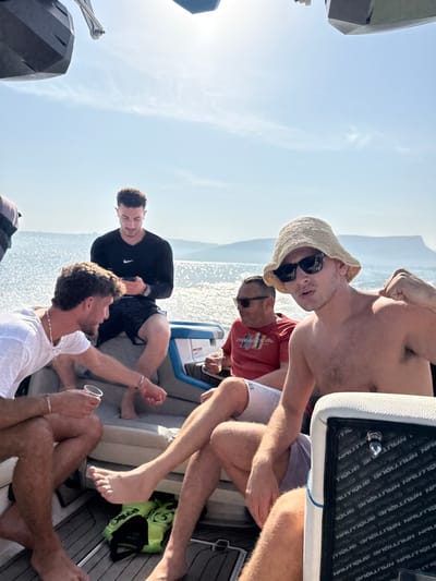 Five adult males are observed on a white and grey modern motorboat on the Sea of Galilee (Lake Kinneret), in the vicinity of Migdal, Israel, under a clear, bright blue sky with direct sunlight reflecting on the water.
The boat features padded seating with grey and light blue accents, and a dark, textured floor where a neon yellow and black life vest rests. From left to right:
1. A man with dark hair, wearing a black long-sleeved Nike shirt and dark patterned shorts, is seated, looking down at a black smartphone held in both hands.
2. A man with curly brown hair and a beard, wearing a white t-shirt and light-colored shorts, holds a clear plastic cup in his left hand and gestures downwards with his right, appearing to interact with something at floor level. He wears silver chain necklaces and bracelets.
3. A man with short dark hair, wearing sunglasses and a red t-shirt with a colorful graphic ("EXPLORE NATURE"), is seated, holding a clear plastic cup or bottle, and looking towards the left.
4. In the foreground, a shirtless man with light hair, wearing sunglasses and a straw bucket hat, is seated, facing the camera directly, and flexing his right arm. He wears light-colored shorts.
5. The legs of a fifth man, wearing shorts, are visible in the extreme foreground on the right.
Most individuals appear barefoot. Distant hills are visible on the horizon across the sparkling water. A "NAUTIQUE" brand label is partially visible on the white side padding of a seat in the foreground. The men appear engaged in leisure activities.