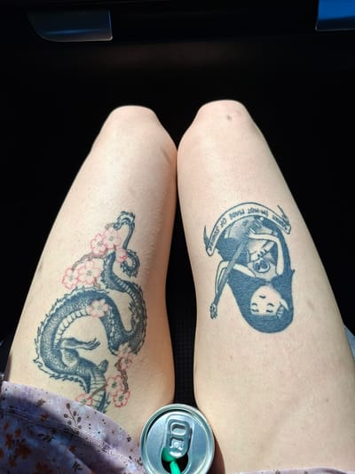 The image shows two human legs with tattoos. On the left leg is a tattoo of a female figure cradling a skeleton while playing a stringed instrument. A banner surrounds the image with the text "Sorry I'm not made of sugar". On the right leg, there is a tattoo of a dragon intertwined with cherry blossom branches. A partially visible can of a drink with a green straw rests next to the right leg. The background is dark, suggesting an indoor setting, possibly a vehicle. The image does not provide any information regarding the location in the city of Kamburovo, Bulgaria.