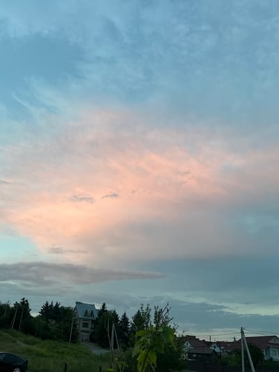 The image depicts a twilight sky with soft pink and blue hues from the setting sun illuminating scattered clouds. Below the sky, a suburban landscape is visible, featuring dense green trees that partially obscure several houses with grey, gabled roofs. A paved driveway leads to one of the houses, and a dark-colored car is parked on the left side of the frame. The scene is set in Новотерский, Russia, as indicated by the location tag.
