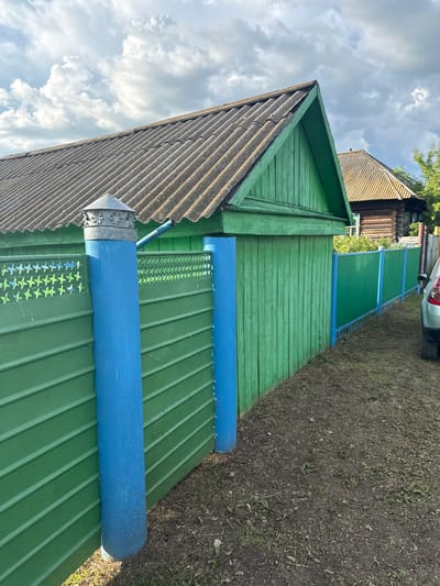 In the village of Chuy-Ataevo, Russia, a weathered, grey, corrugated roof caps a rustic green wooden structure. A blue cylindrical post, painted in a bright, matte finish, supports the structure and a section of green wooden fence. This fence, constructed from horizontal planks, is adorned with a decorative border of light green and blue star-like patterns along its top edge. Beyond this fence, a secondary green corrugated metal fence with blue posts stretches into the distance, bordering a grassy area. In the background, another wooden building with a similar grey roof is visible, along with lush green foliage. The sky above is a dramatic mix of light and dark grey clouds. The ground in the foreground is a mixture of sparse grass and dark soil. A portion of a silver car is visible on the far right of the image.