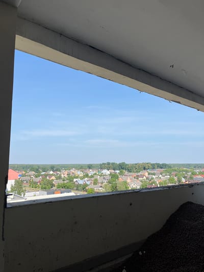 The image depicts a view of a village from a partially constructed building in Аснежыцкі сельскі Савет, Belarus. The foreground shows a concrete ledge and a pile of dark, small, round objects, possibly gravel or construction debris. Beyond this, a bright blue sky with faint clouds stretches across the horizon. Below the sky lies a dense arrangement of residential houses with red, grey, and yellow roofs, interspersed with green trees. A dark green line of forest borders the village in the distance. The overall impression is of an undeveloped area with ongoing construction.