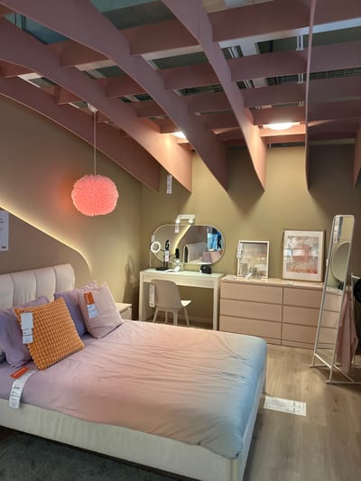 The image depicts a bedroom staged in a retail environment, likely an IKEA store, given the visible price tags and product display. The room features a bed with light-colored bedding, adorned with purple and orange throw pillows. A vanity table with a mirror and lighting is situated against the wall, accompanied by a chair. To the right, a dresser and a full-length mirror are visible. The ceiling has a distinctive pink architectural element with exposed beams and lighting fixtures. The walls are painted a neutral taupe color. The floor is wooden laminate. No people are explicitly depicted in the scene, but reflections in the mirror suggest other shoppers may be present in the background. The overall aesthetic is modern and feminine, with a color palette of pastels and neutrals. The location is identified as an IKEA store in Siero, Spain.