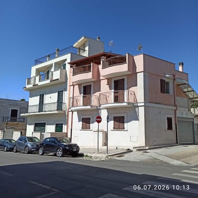 A full-shot, eye-level, medium-wide outdoor photograph captures a clear, bright, sunny day in Ruvo di Puglia, Italy, with a deep blue sky overhead. The scene is dominated by a modern, multi-story building with a light pink facade on the right and a white facade on the left, featuring balconies with dark railings on multiple levels. The building has brown shutters on its windows and several chimneys and satellite dishes on the roof. Adjacent to the main building on the left is a lighter colored stone building with a dark brown shuttered window and a balcony. In front of the buildings, parked along the street, are three cars: a dark sedan on the left, a gray SUV in the middle, and a black SUV on the right. A circular red and white "no entry" sign is mounted on a pole in front of the pink building. The street in the foreground is paved with asphalt and a pedestrian crosswalk is visible at the bottom of the frame. A timestamp in the lower right corner reads "06.07.2026 10:13".