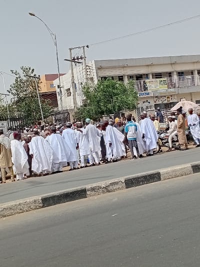 The image displays a daytime street scene in Sani Mai Nagge, Nigeria. In the foreground, an asphalt road is bordered by a concrete curb featuring a black painted upper edge. A large group of individuals, predominantly male, stand on the pavement adjacent to the curb. Most are dressed in long white robes and traditional caps, while a few wear contemporary clothing. A motorcycle is partially visible among the crowd on the right side. In the mid-ground, a multi-story building, likely commercial, displays signs such as "AGIANA HOTEL," "ALBARKA STYLE," and "ZAM ZAM." Further back, additional buildings, green trees, a tall street light, and utility poles with overhead power lines are visible under a bright, clear sky. The crowd appears to be stationary, facing generally towards the right of the frame.