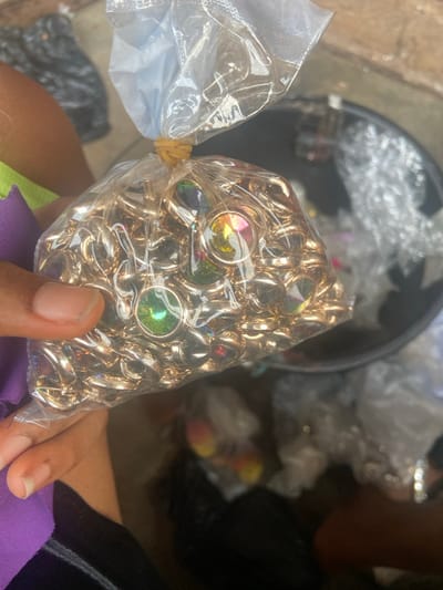 A clear plastic bag tightly holds a large quantity of gold-colored metal rings. Each ring has a centrally placed, iridescent jewel that reflects various colors. The bag is secured with a yellow rubber band. An unidentifiable purple fabric lies underneath the bag being held by a person's hand, with only the fingers and part of the palm visible. Other bags filled with similar rings are seen in the background, stored in a black container.