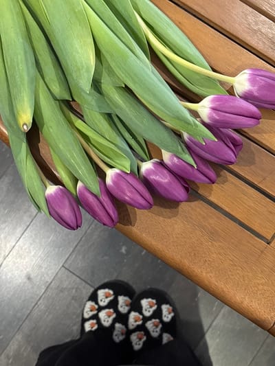 The image displays an indoor scene in Trudobelikovskiy, Russia. A bouquet of approximately twelve purple tulips, characterized by vibrant green stems and leaves and closed purple flower heads, is laid horizontally across a light brown, slatted wooden surface. Below and to the left of this wooden surface, the feet of an individual are visible, wearing black socks patterned with multiple white cartoon ghost figures featuring small orange noses or mouths. The feet are positioned on a dark gray floor, which appears to be either laminate or tiled.
