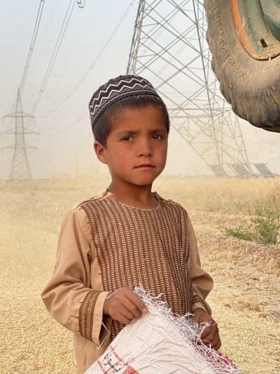 A young boy stands in a field in Alaqadari Daman, Afghanistan, holding a white sack. He wears a tan tunic with brown embroidered trim and a patterned kufi hat. Behind him, electrical towers are visible, and a large tractor tire is partially in the frame on the right. The ground appears to be covered in harvested grains. The air seems dusty.
