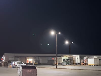 The image depicts an industrial area, likely a warehouse or distribution center, located in Plodovitovo, Bulgaria at night. The scene is illuminated by streetlights, with a long, low building forming the backdrop. Several delivery trucks and forklifts are parked near loading docks. A white van is parked in front of one of the open loading bays, with boxes and a person nearby. A large dumpster sits in the foreground. Stop signs are visible near the building. The sky is dark, with faint green artifacts possibly from camera.