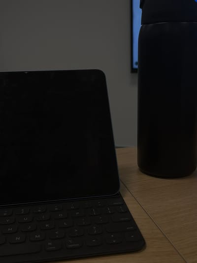 A black water bottle sits on a wooden surface, partially overlapping a black keyboard and tablet. The tablet's screen is dark, and the keyboard's keys are visible, including letters like "Y," "U," "K," "J," "H," "N," and "M." The keyboard also has keys labeled "option" and "cond." A faint blue light is visible in the background, possibly emanating from a screen.