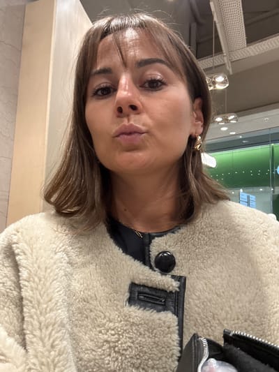 A woman with brown hair and bangs is pictured close-up. She has fair skin, brown eyes, and is making a pursed-lips expression. She wears a gold earring and a tan sherpa jacket with black leather trim. Part of a black leather handbag is visible. The background includes blurred lights and a greenish tint. A beige surface is in the lower left corner.