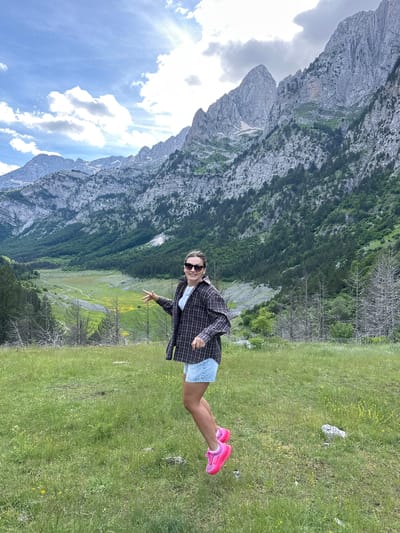 A woman stands in a grassy field with a scenic mountainous backdrop. The woman is wearing a plaid shirt, denim shorts, and bright pink athletic shoes. She is smiling and gesturing with her right hand, her left arm bent with her hand resting on her hip. Her sunglasses are on her face. The landscape behind her features rolling green hills that lead to jagged, rocky mountains. Sparse trees dot the foreground and midground, with some appearing dead and bare. The sky above is blue with scattered clouds and sunlight filtering through. The image appears to be taken in the vicinity of Shalë, Albania, a location known for its dramatic mountain scenery.